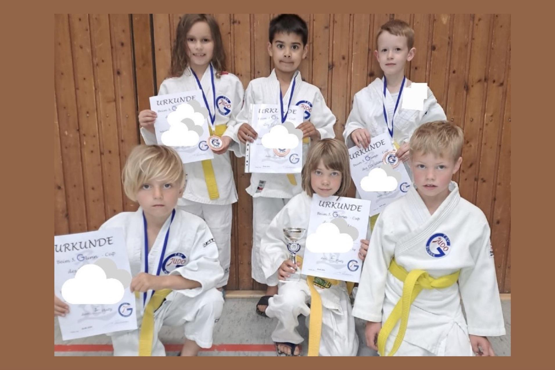 Gruppe von sechs Kindern in Karateanzügen mit Medaillen und Urkunden vor einer Holzwand.