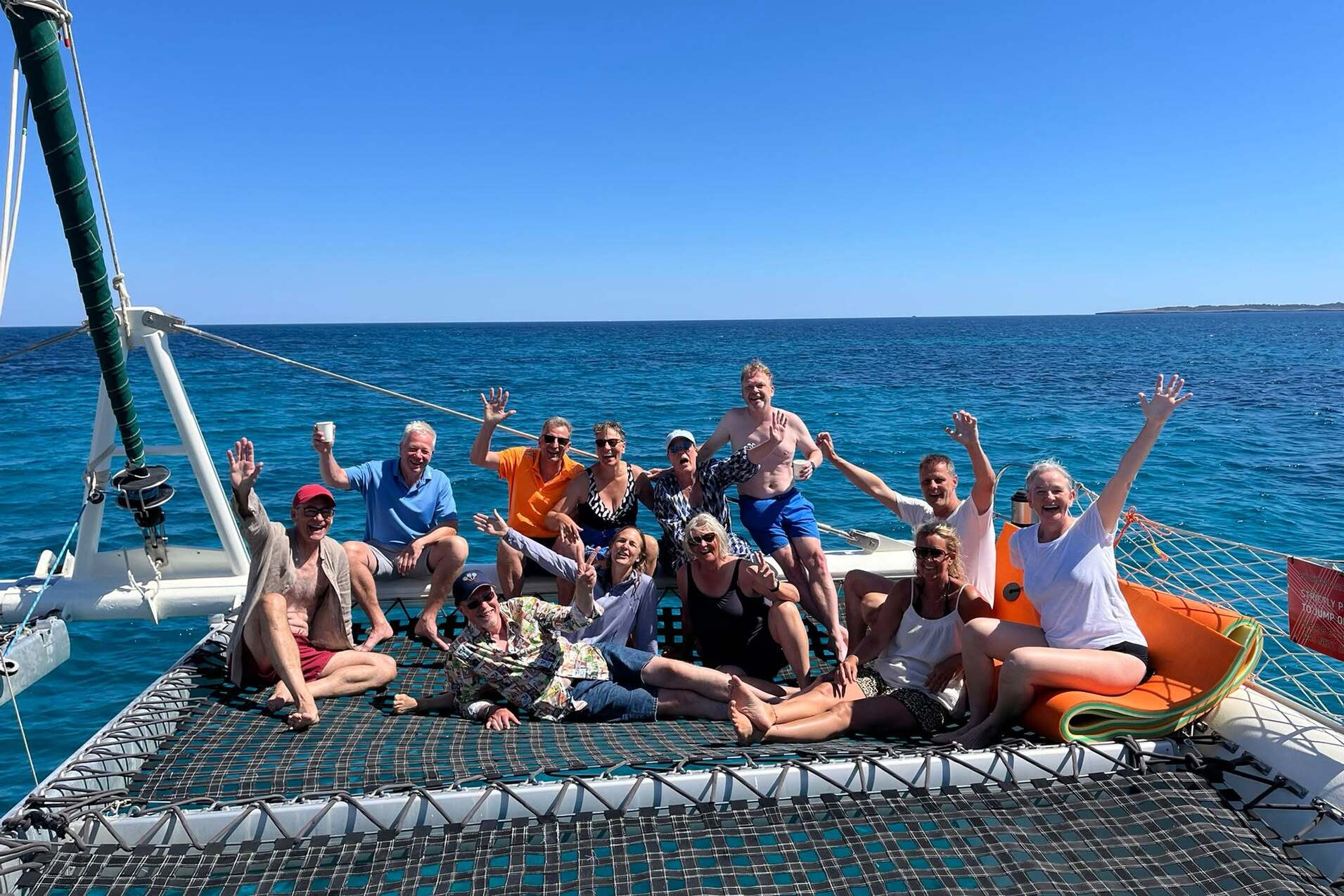 Gruppe von 15 Personen auf einem Katamaran-Netz, lächelnd und winkend, mit blauem Meer im Hintergrund.